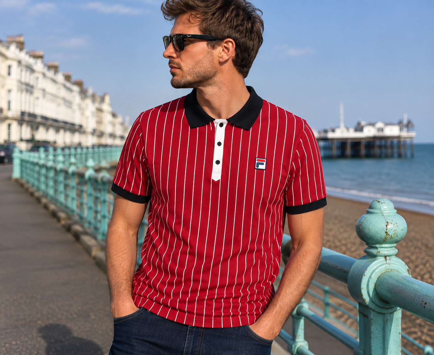 A man wearing a red and white vertically striped polo shirt with a black collar and black trim stands on a seaside promenade with a pier and ocean in the background. A small Fila logo is visible on the chest of the shirt.