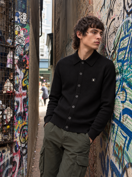 A man wearing a black knit polo cardigan and olive green cargo pants stands in a narrow urban alleyway with graffiti-covered walls.