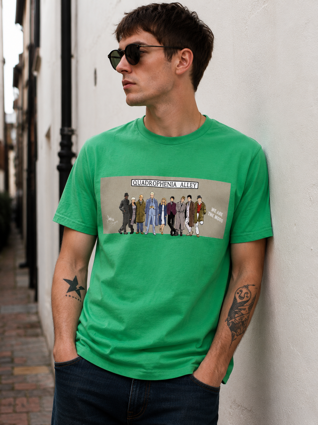 A man wearing a bright green graphic t-shirt with a 'Overground' band print, standing against a white wall.